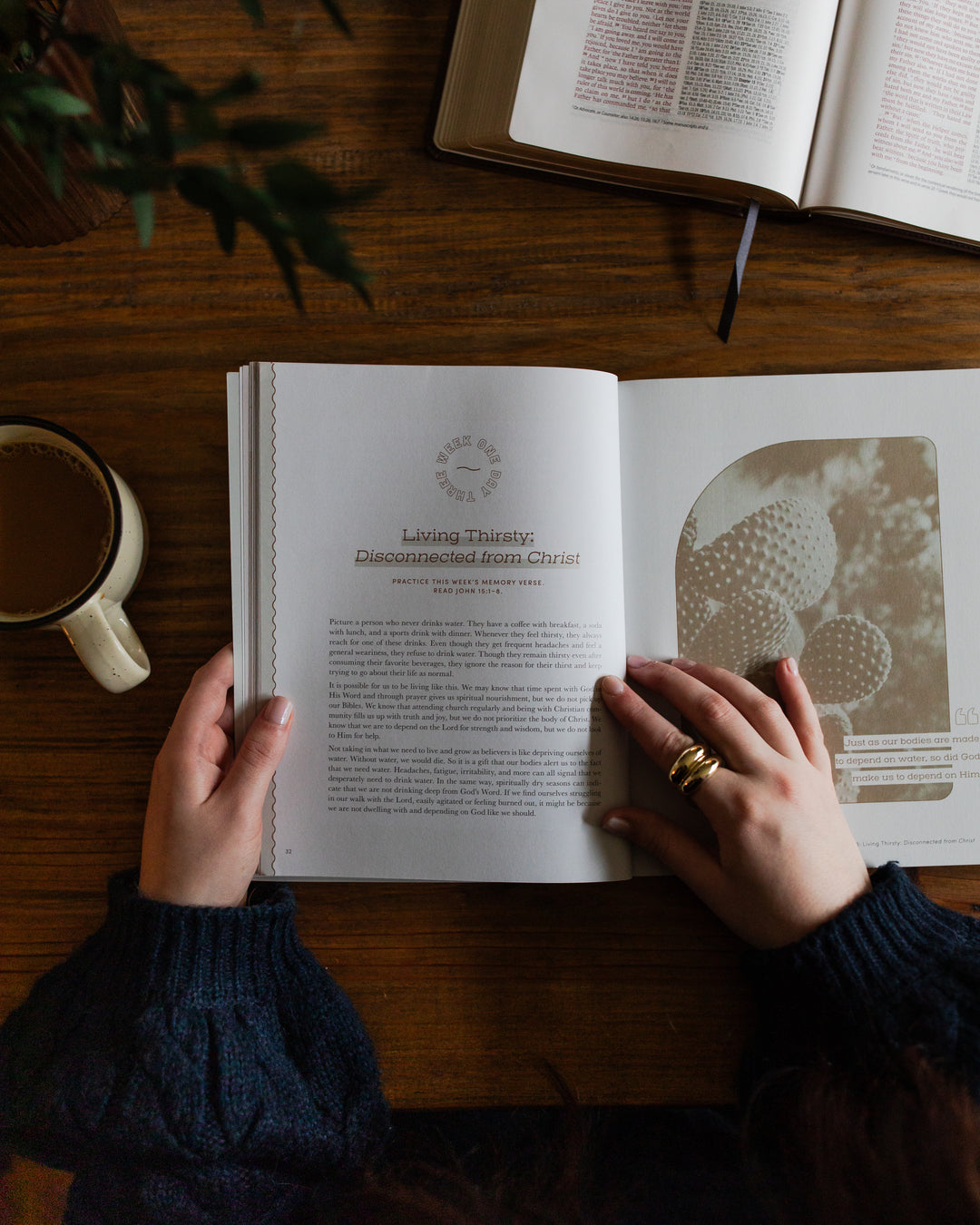 Rivers of Renewal Study opened to week one day three page laying on a brown wooden table; A woman is holding the study open with one hand in the middle of the page; There is a coffee cup and an open bible laying on the table next to the study