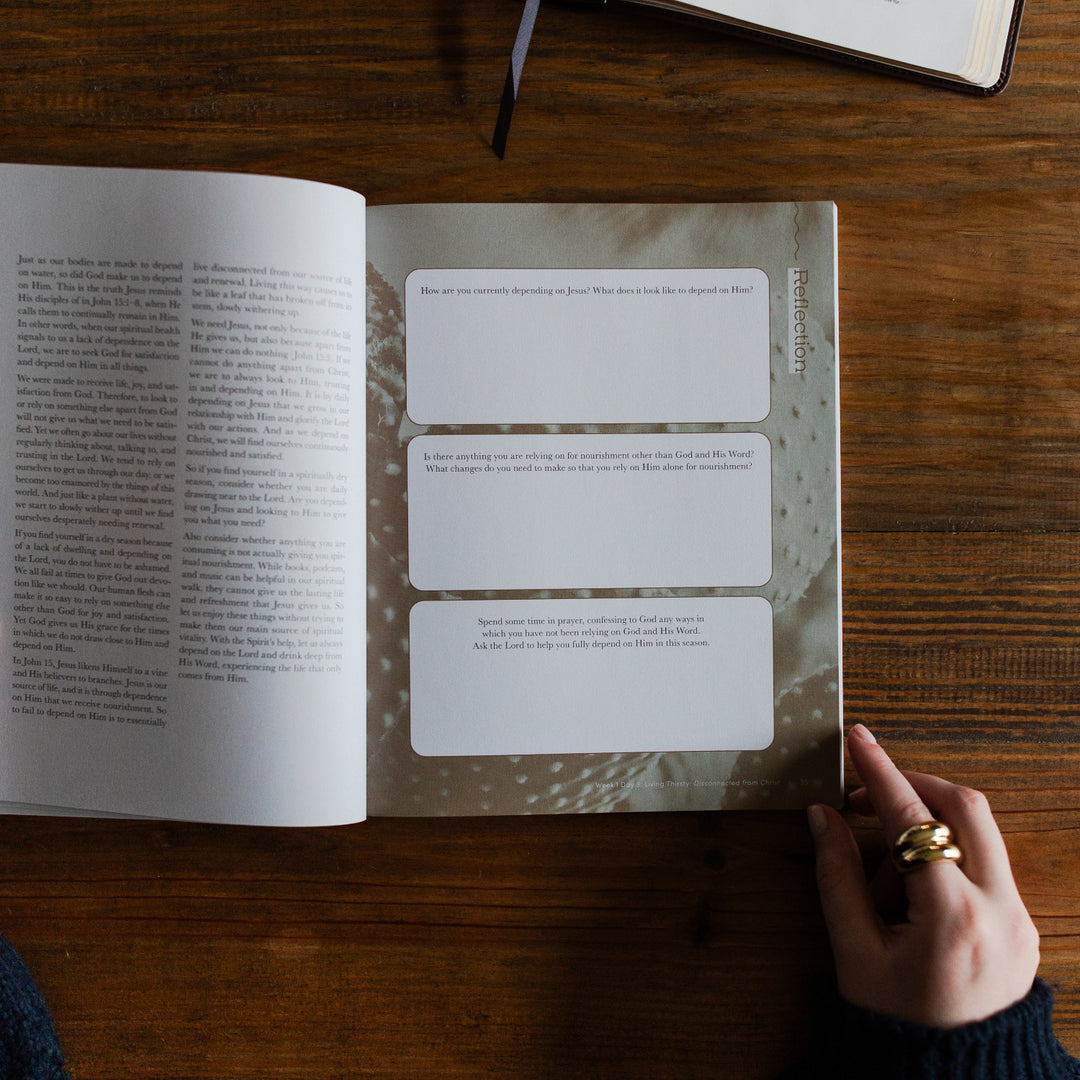 Rivers of Renewal Study opened to a reflection page laying on a brown wooden table; A woman is holding the study open; There is an open bible laying on the table next to the study