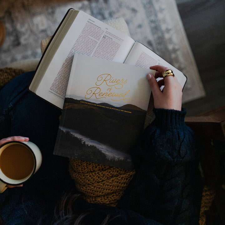 Rivers of Renewal Study resting on an open bible; a woman is holding the corner of the study and a cup of coffee while the bible and study rest on the arm of a couch.