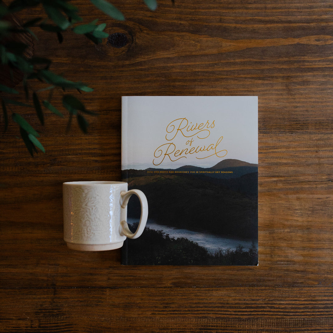 River of Renewal Woman's study cover next to a Daily Grace Mug laying on a brown wooden table with greenery in the corner