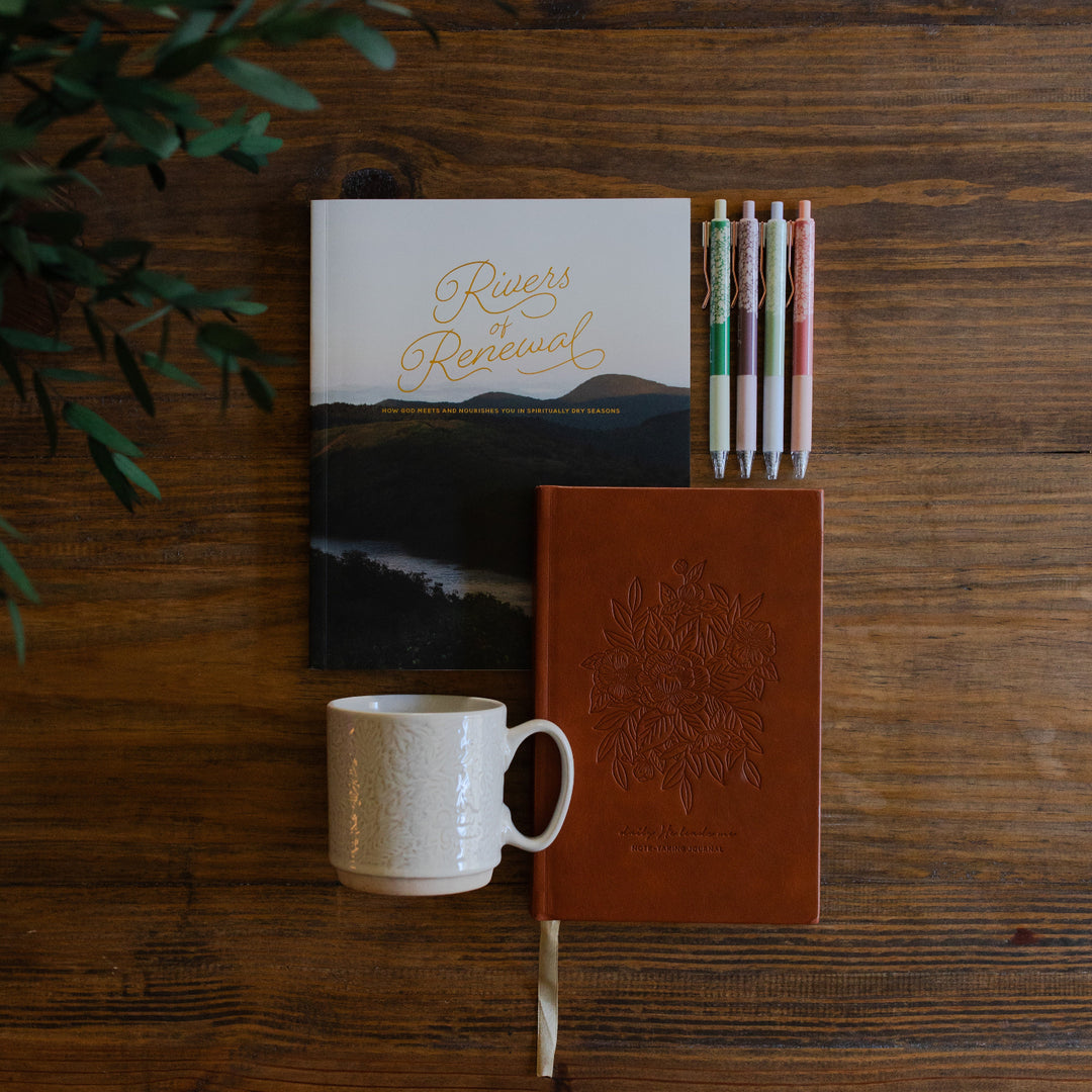 River of Renewal Woman's study cover next to a He leads me Journal, a set of Psalm 23 pens, and a Daily Grace Mug laying on a brown wooden table with greenery in the corner