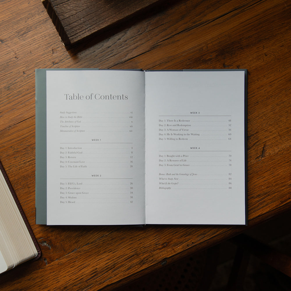 An open book laid flat on a rustic wooden table, showing the Table of Contents spread across both pages. The book has a muted green-gray cover and white interior pages with black text.