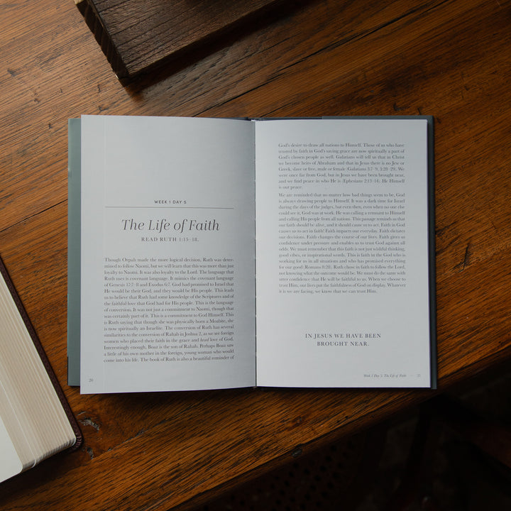 An open book laid flat on a wooden table, showing two printed pages with text from 'The Life of Faith'. The book has a gray cover and is surrounded by a dark wooden surface with other books partially visible at the edges.