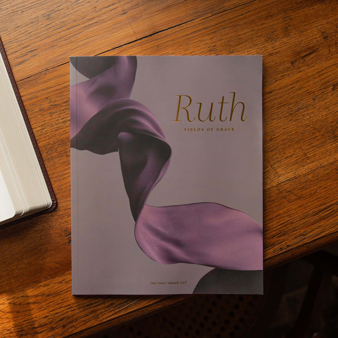 A book titled 'Ruth: Fields of Grace' by The The Daily Grace Co. lies on a warm wooden table. The visible cover features a flowing purple ribbon design on a muted lavender-gray background.