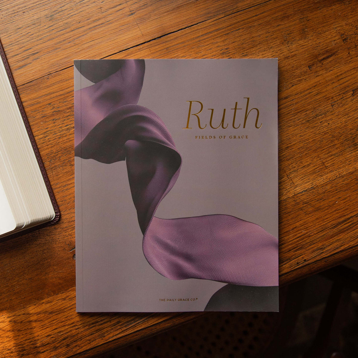 A book titled 'Ruth: Fields of Grace' by The The Daily Grace Co. lies on a warm wooden table. The visible cover features a flowing purple ribbon design on a muted lavender-gray background.