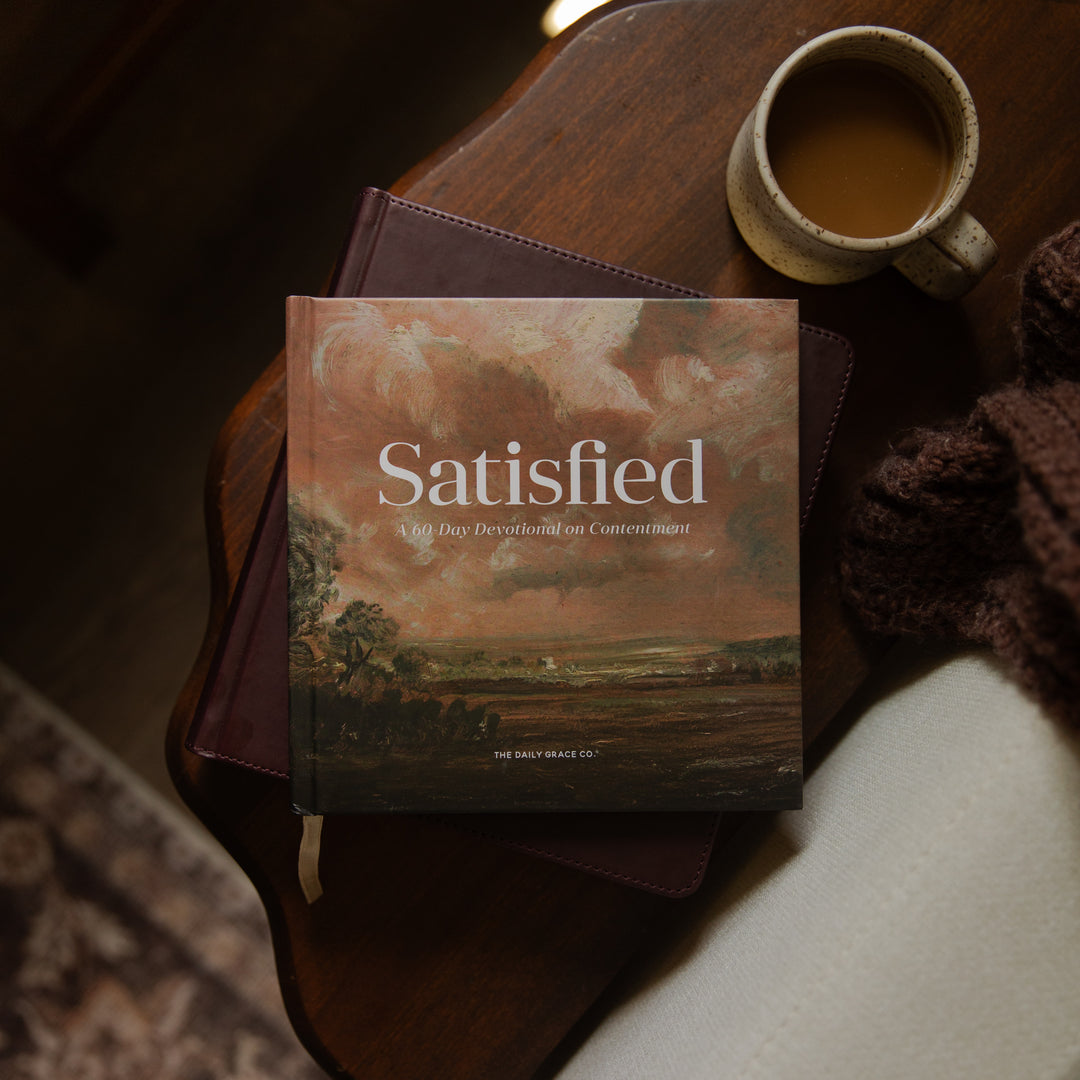 A book titled "Satisfied" by The Great Sage Co. lies on a dark wooden table next to a ceramic mug of coffee and a knitted brown scarf.