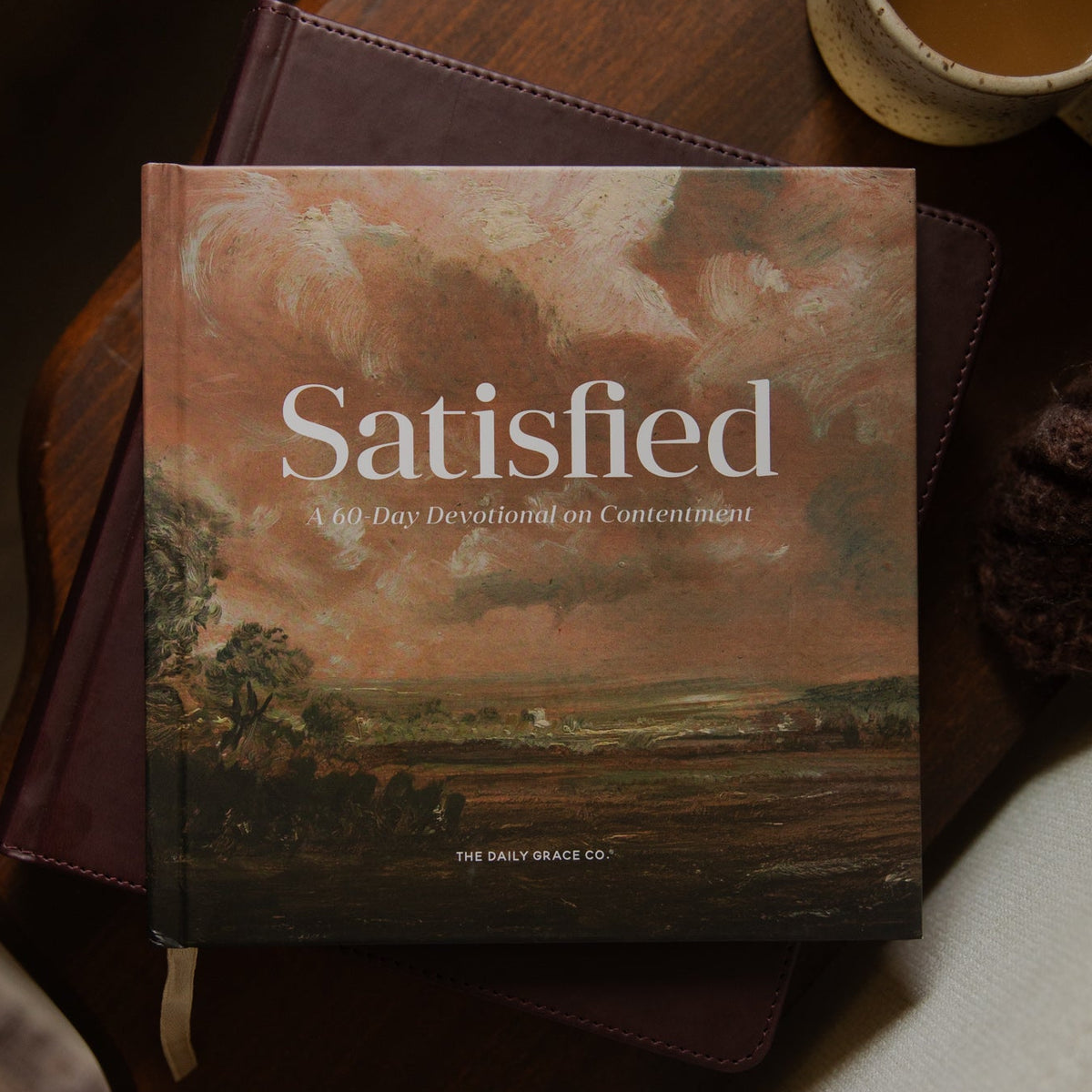 A book titled "Satisfied" by The Great Sage Co. lies on a dark wooden table next to a ceramic mug of coffee and a knitted brown scarf.