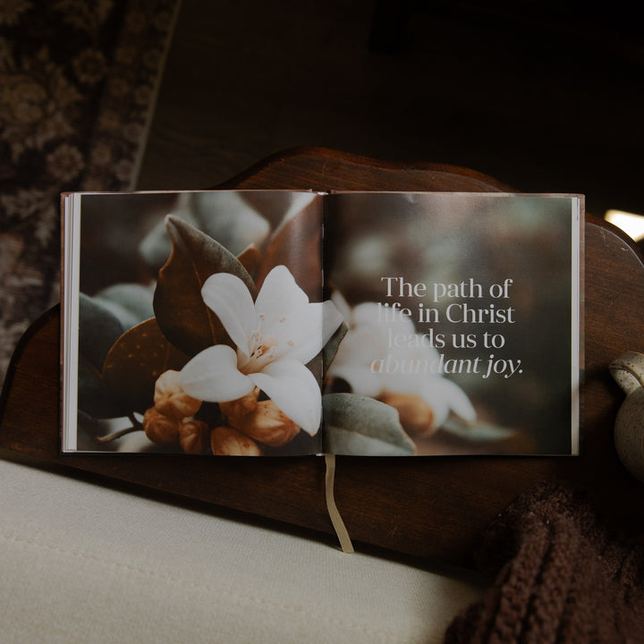 An open printed book or journal resting on a dark wooden surface, showing a floral photograph on one page and the quote, 'The path of life in Christ leads us to abundant joy,' on the other. A cream ribbon bookmark hangs from the center.