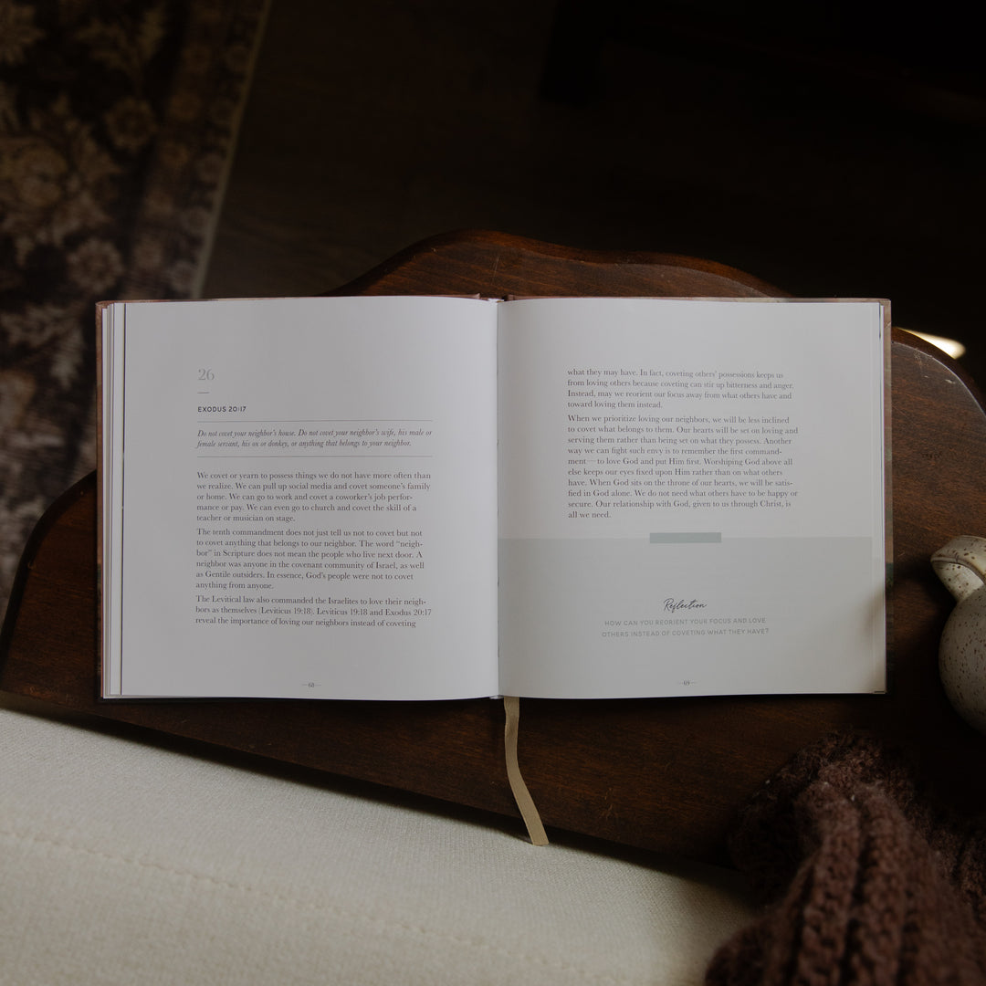 An open book with printed text spread across two pages, resting on a dark wooden surface with a cream fabric and brown knit textile nearby.