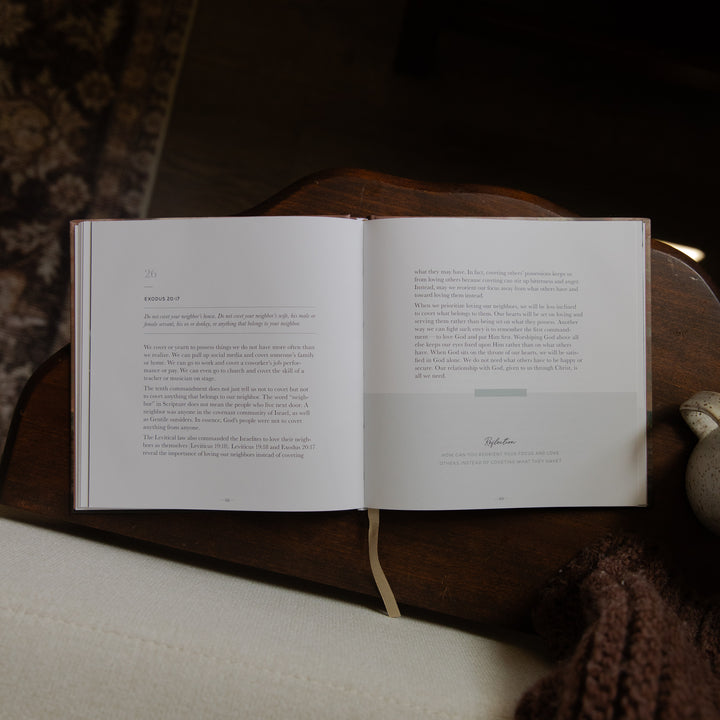 An open book with printed text spread across two pages, resting on a dark wooden surface with a cream fabric and brown knit textile nearby.