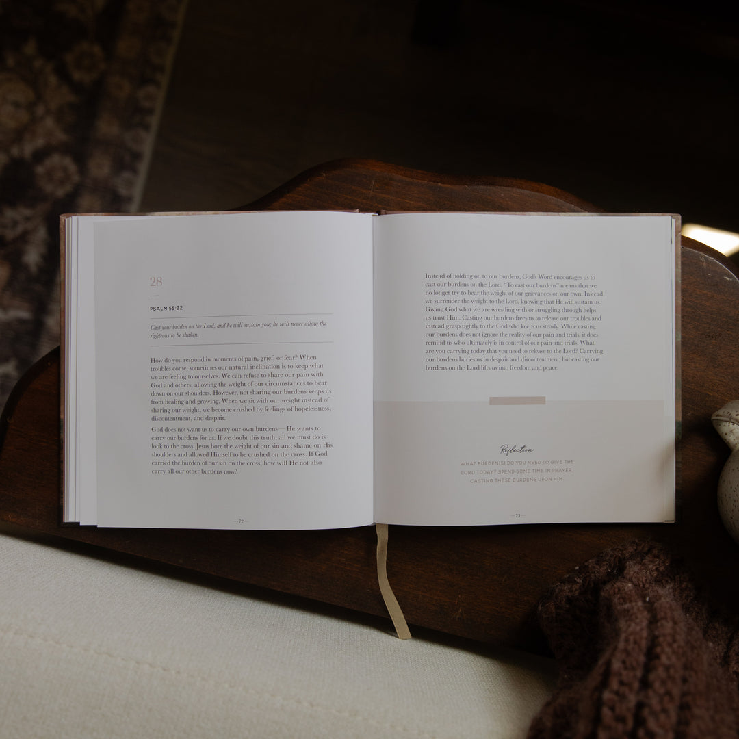 An open book with printed text spread across two pages, resting on a dark wooden surface with a cream fabric surface below and a brown knit textile at the lower right.