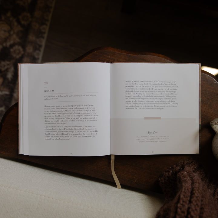 An open book with printed text spread across two pages, resting on a dark wooden surface with a cream fabric surface below and a brown knit textile at the lower right.