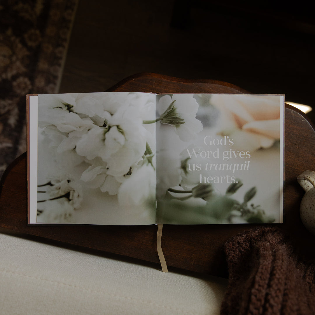 An open book or journal lying on a dark wooden surface, featuring floral photography with the text 'God's Word gives us tranquil hearts.' A cream ribbon bookmark hangs from the center.