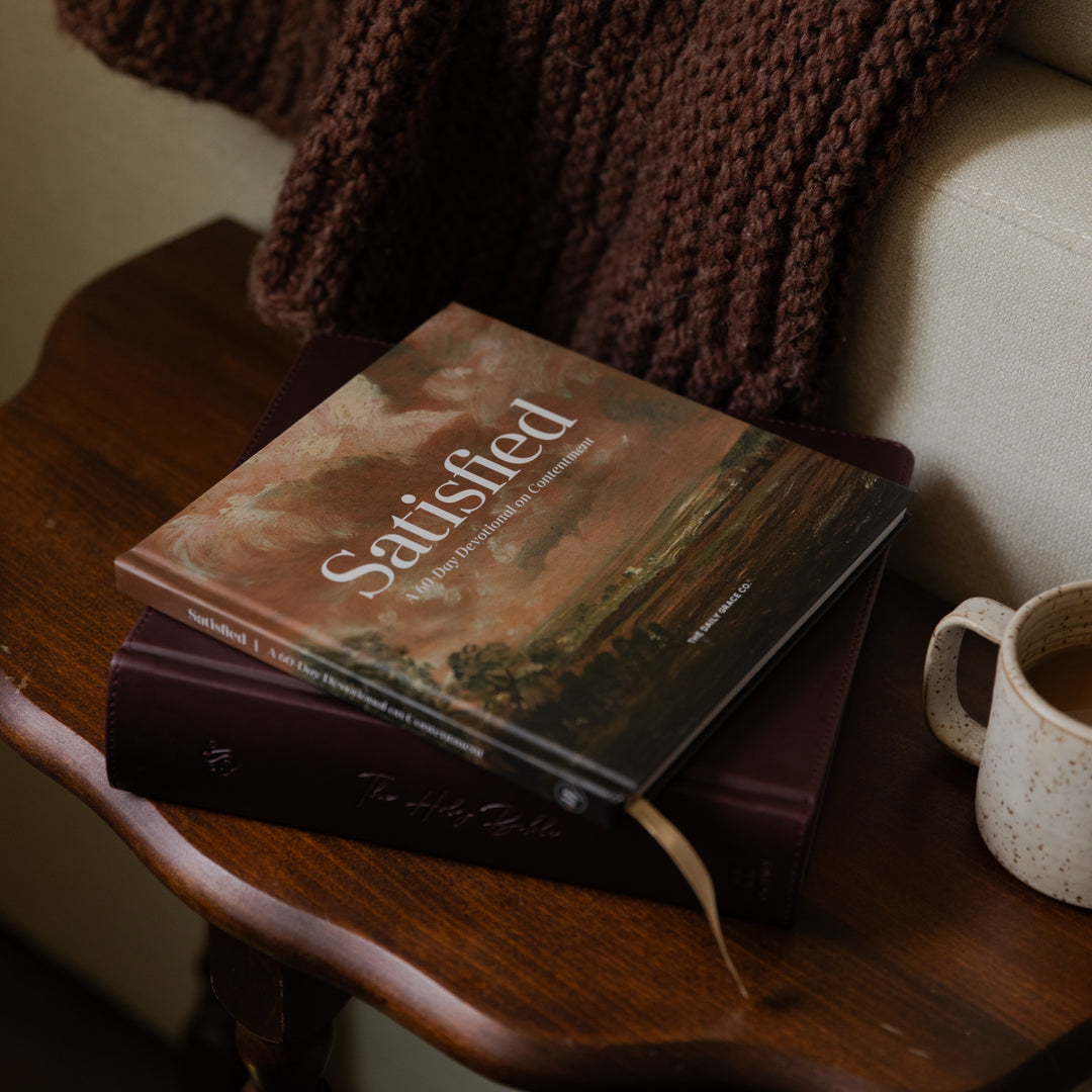 A book titled 'Satisfied' with a landscape cover sits on top of a dark burgundy book on a wooden table, with a chunky brown knit blanket draped over the arm and a speckled mug nearby.