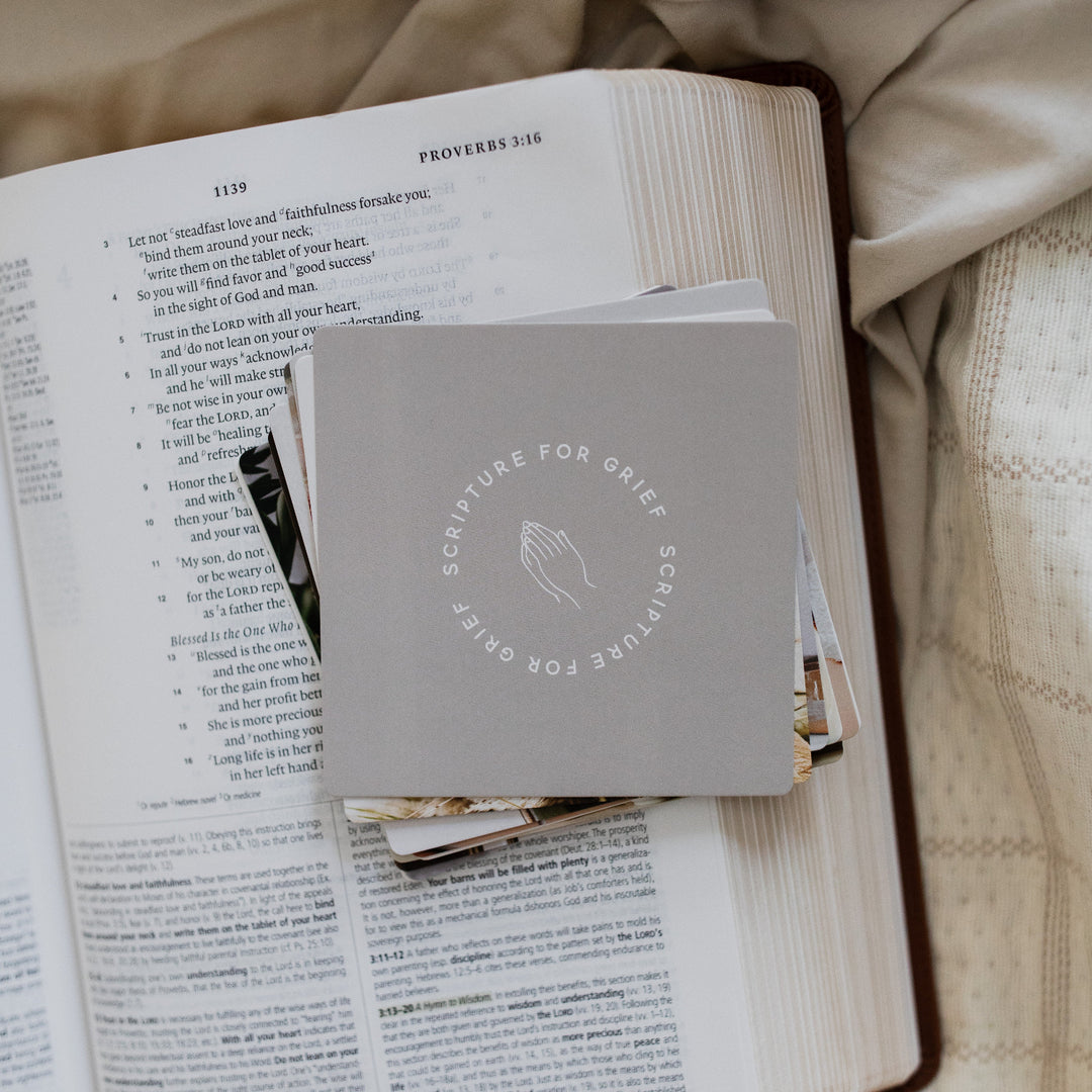 A stack of Scripture for Grief Verse Cards sitting on an open bible; The bible is resting on a a light tan colored cloth