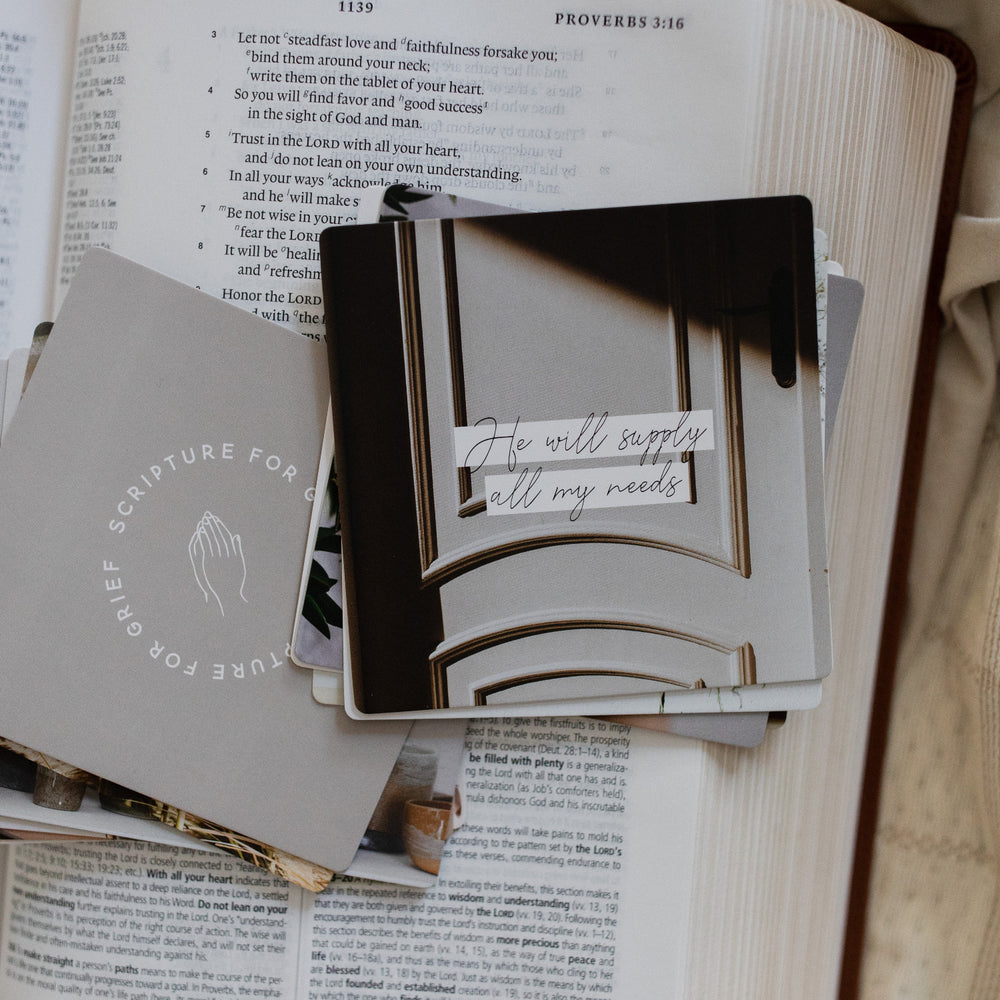 A stack of Scripture for Grief Verse Cards split in two stack sitting on an open bible; The bible is resting on a a light tan colored cloth