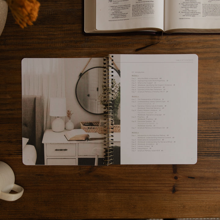 An open spiral-bound journal or planner lies on a wooden table, showing a bedroom photo on the left page and a list of verses from Psalm 34 on the right page. An open book and part of a mug are visible around it in a styled flat-lay setting.