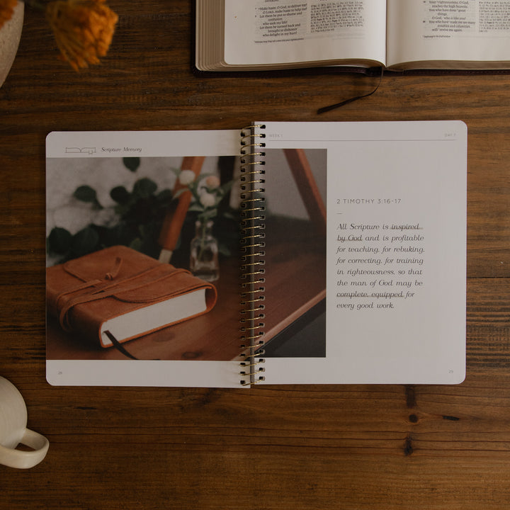 An open spiral-bound journal or notebook lying on a wooden desk, showing a page with a photo of a leather-bound book and a Bible verse from 2 Timothy 3:16-17. Other books and a cup are partially visible around it on the desk.