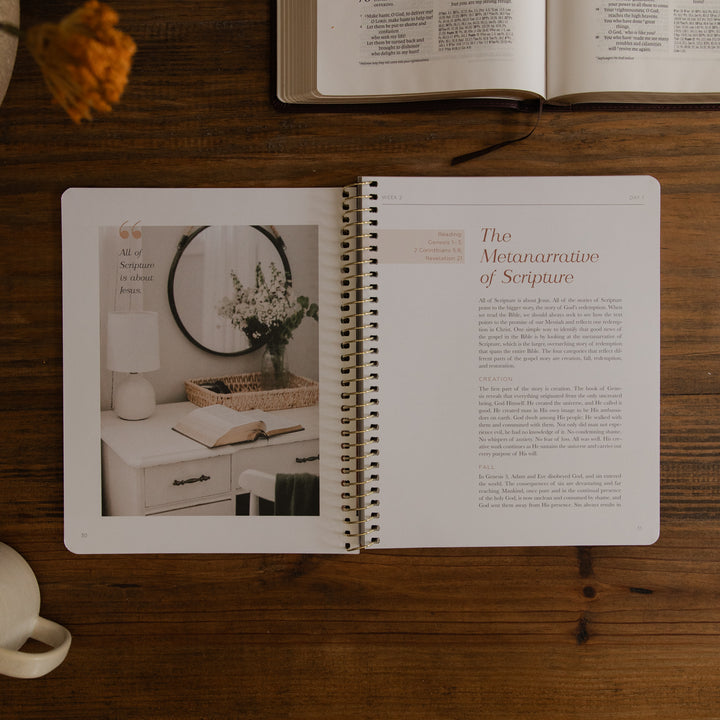 An open spiral-bound book or journal lies on a wooden tabletop, showing a page titled 'The Metanarrative of Scripture' with text and a left-page photo of a styled interior scene. An open book and a small vase are partially visible around it.