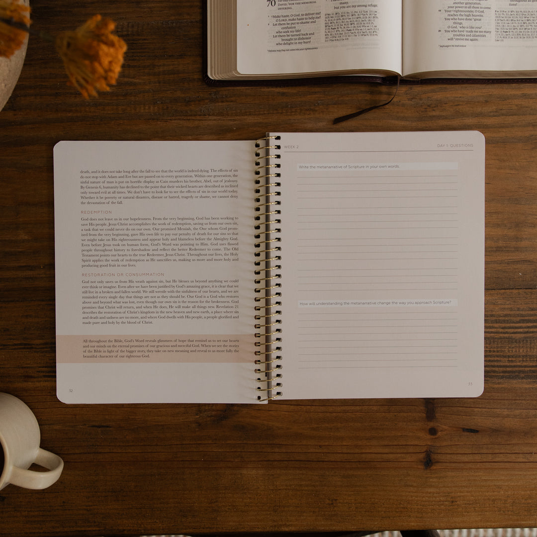 An open spiral-bound journal or planner lies on a wooden table, showing lined pages with highlighted text. An open book, a coffee cup, and a dried orange flower are partially visible around it in a styled tabletop setting.