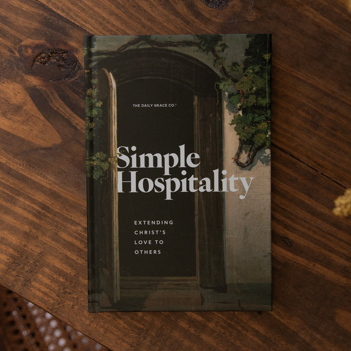 A book titled "Simple Hospitality" lies on a rustic wooden table, surrounded by blurred dried flowers and part of a woven chair. The cover shows a dark arched doorway with green ivy and white title text.