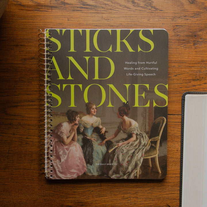 A spiral-bound book titled 'Sticks and Stones: Healing from Harmful Words and Cultivating Life-Giving Speech' lies on a wooden tabletop, styled with a ceramic vase and an open Bible nearby.