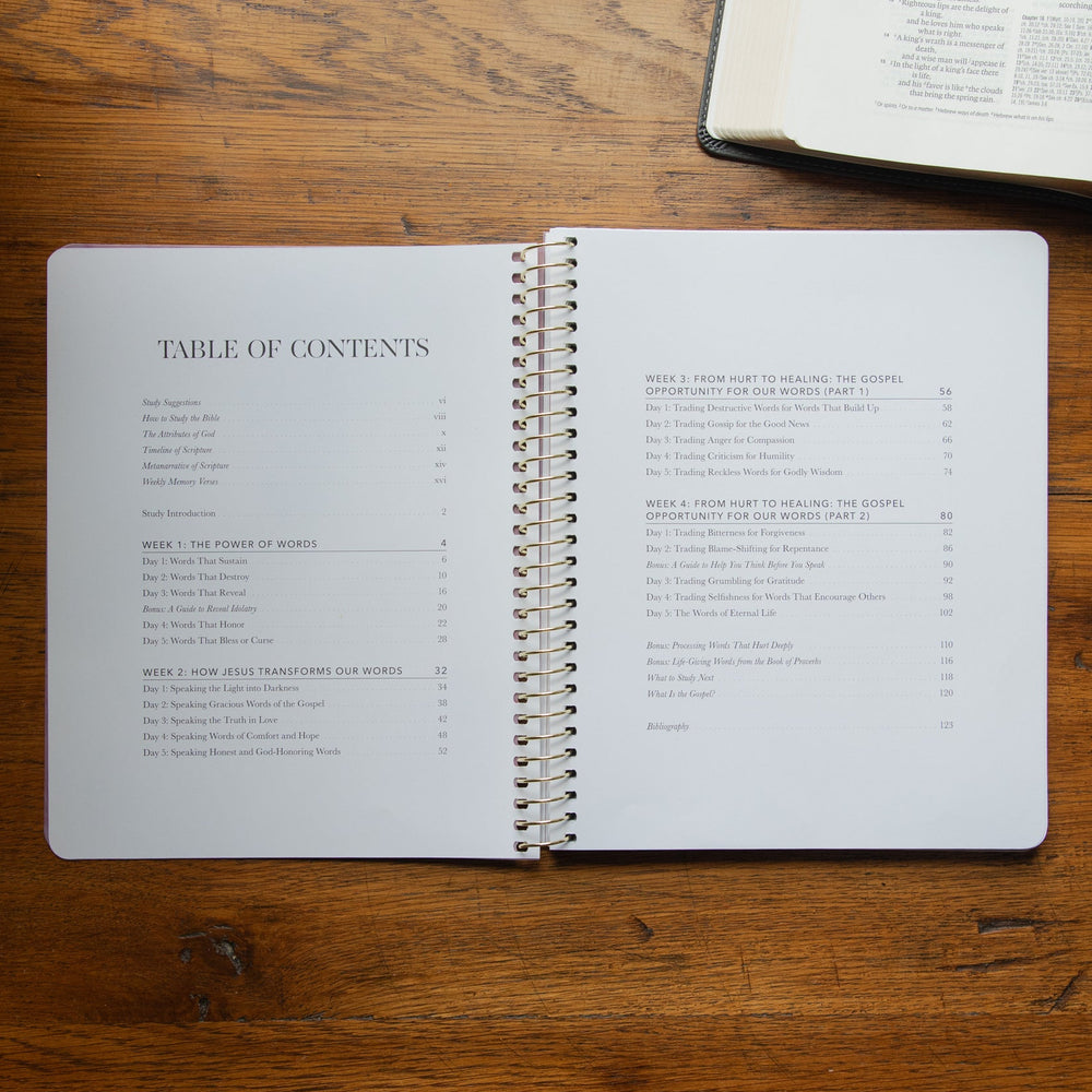 An open spiral-bound book showing a table of contents on a warm wooden tabletop, with another open book partially visible in the upper right corner.