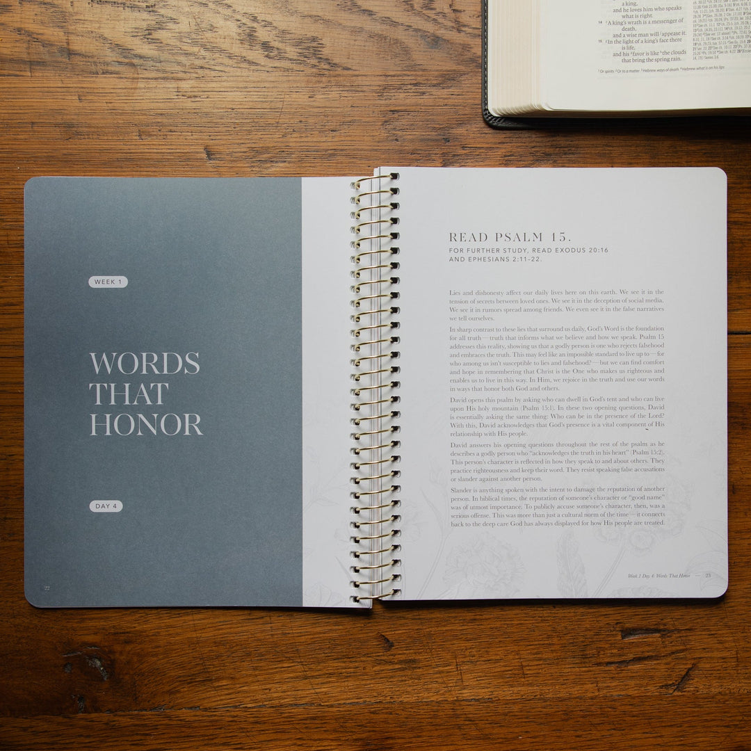 An open spiral-bound book titled 'WORDS THAT HONOR' lies on a wooden table, with a Bible open in the upper right corner. The book has a muted blue-gray cover on the left and printed text pages on the right.