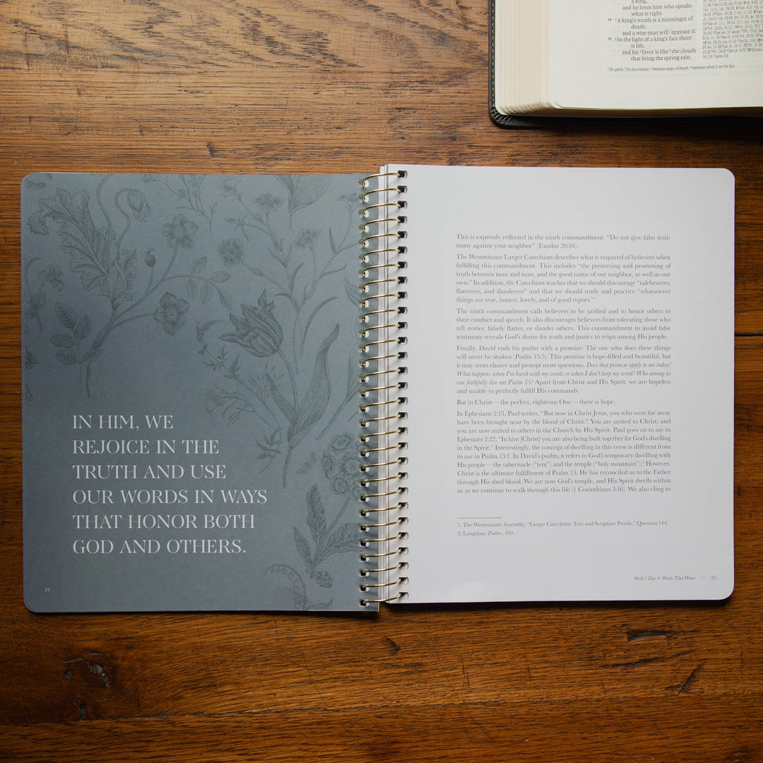 An open spiral-bound study lies on a wooden tabletop, with the left page featuring a muted gray floral border and printed scripture text, and the right page showing text columns. Another open book is partially visible at the top right.