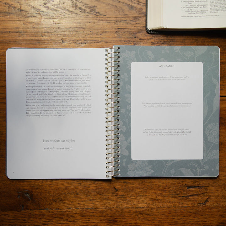 An open spiral-bound booklet set lies on a wooden tabletop, with one page showing printed text and the other featuring a large blank white card labeled 'APPLICATION.' A second open book is partially visible in the top right corner.