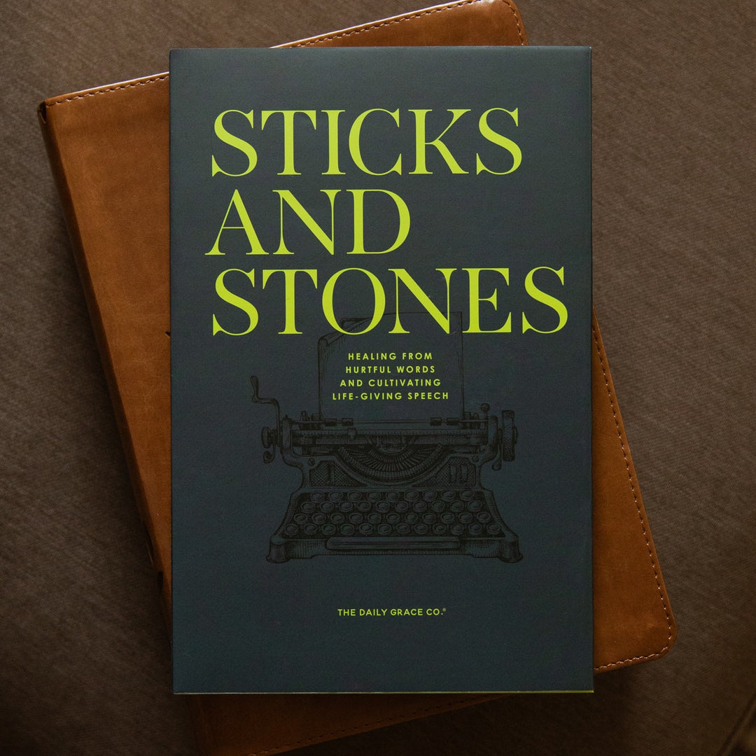 A book titled 'Sticks and Stones' with a dark slate-blue cover and bright yellow-green title text, resting on top of a brown leather-bound book on a brown upholstered surface.