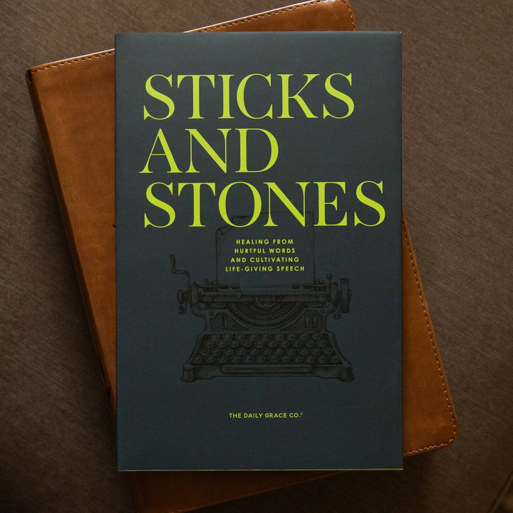 A book titled 'Sticks and Stones' with a dark slate-blue cover and bright yellow-green title text, resting on top of a brown leather-bound book on a brown upholstered surface.