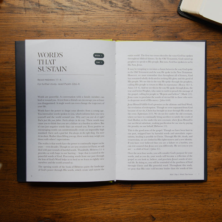 An open book showing printed text on two pages, placed on a wooden tabletop with another book partially visible in the top right corner.