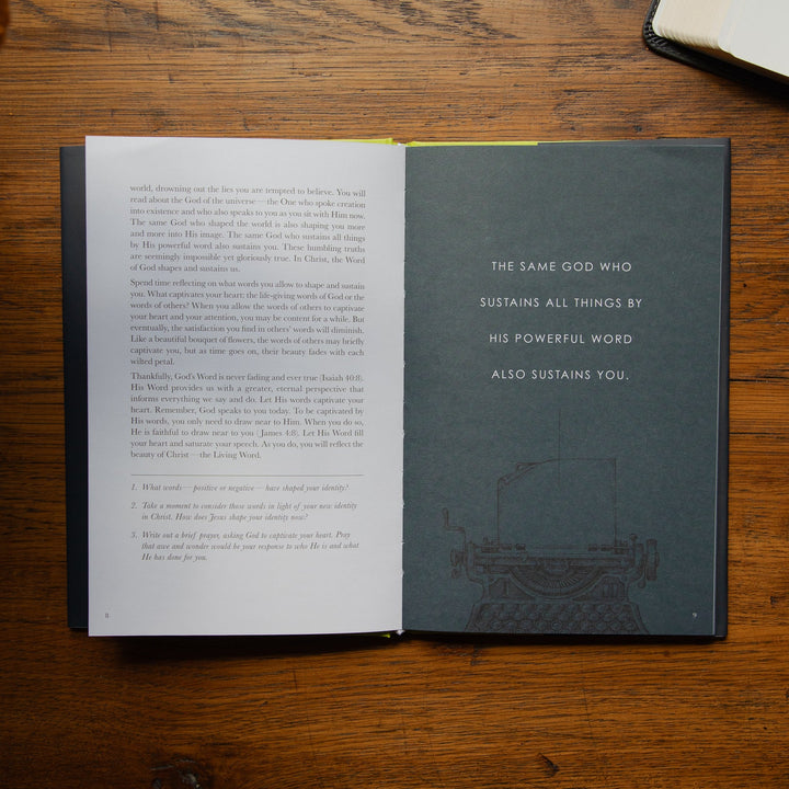 An open book laid flat on a wooden table, showing a two-page spread with black text on white paper and a dark gray right page with a quote and faint illustration. Another open book and dried leaves are partially visible around it.