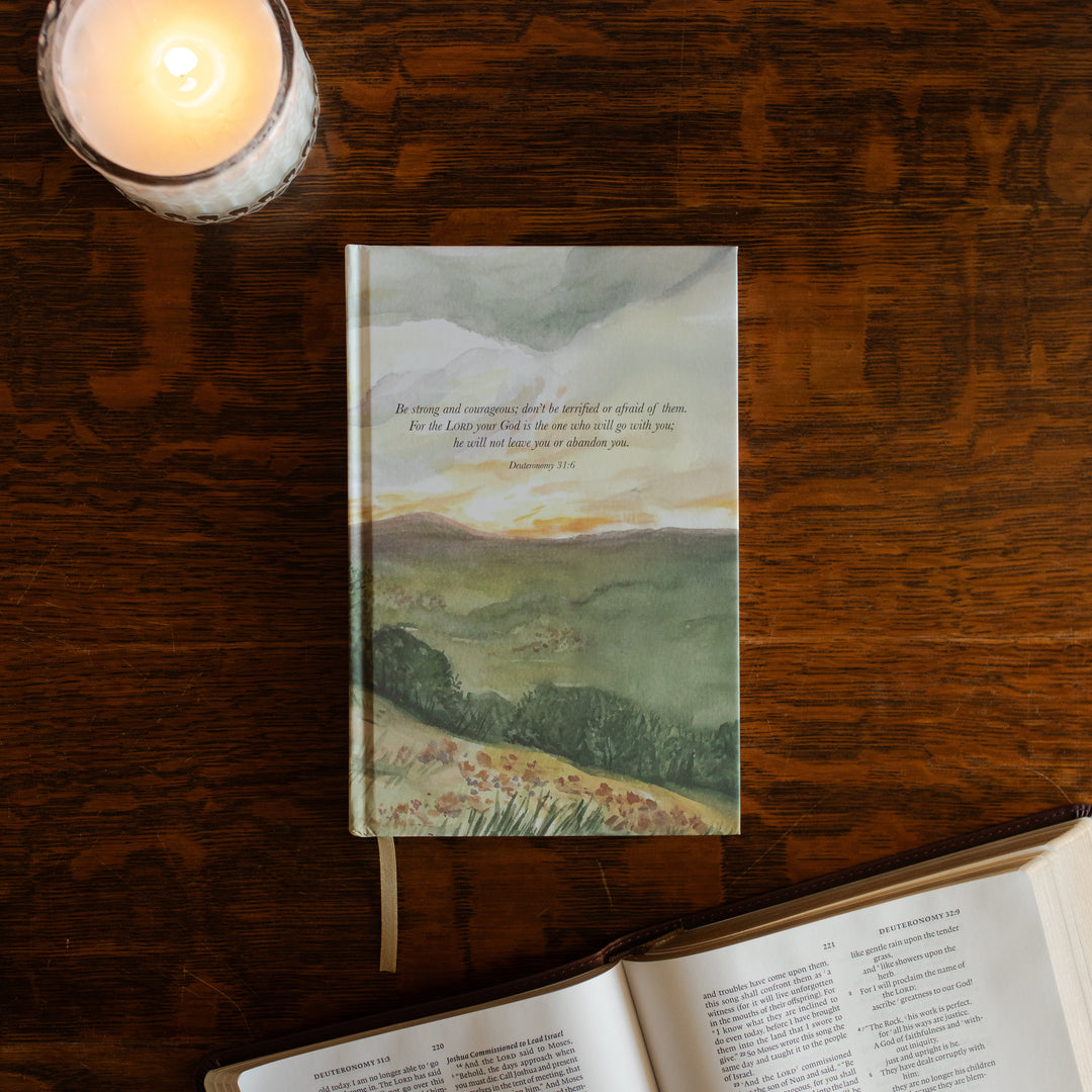 Strong and Courageous Lined Journal cover on a wooden table next to a lit candle and an open bible