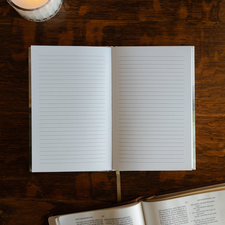 Strong and Courageous Lined Journal opened to a blank page on a wooden table next to a lit candle and an open bible