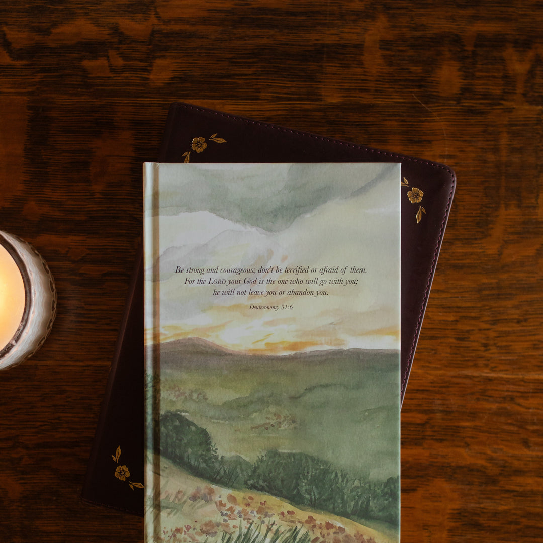 Up close of the Strong and Courageous Lined Journal cover on a closed bible on a wooden table next to a lit candle