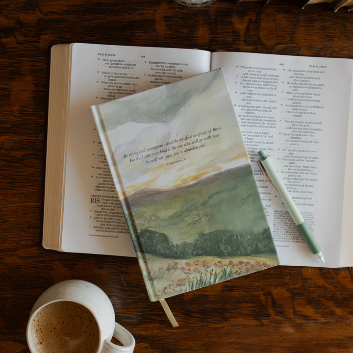 Strong and Courageous Lined Journal cover on an open bible on a wooden table next to a coffee cup