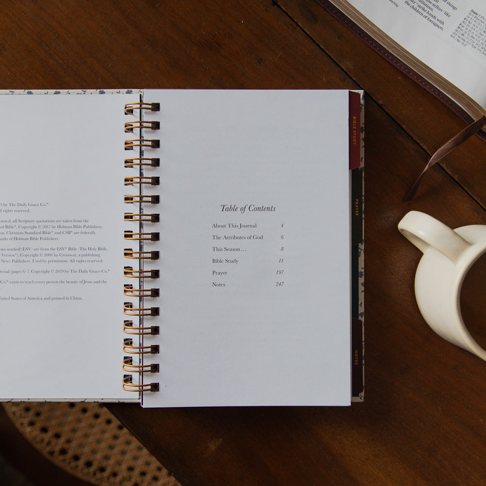 An open spiral-bound journal or notebook showing a table of contents page, placed on a wooden table next to an open book and a white ceramic mug.