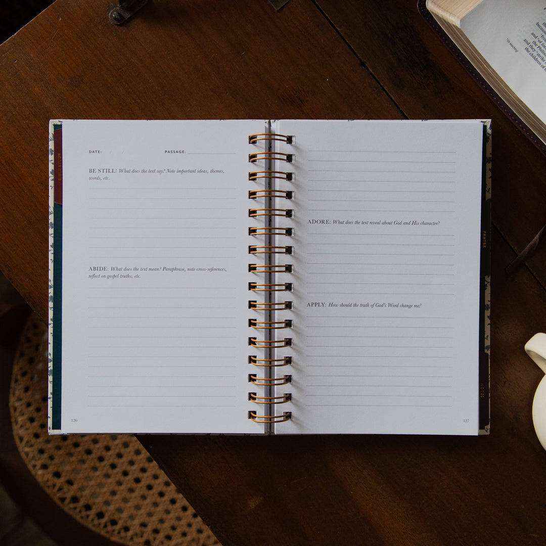 An open spiral-bound journal or planner spread on a dark wooden table, showing lined pages with prompts and a decorative spine.