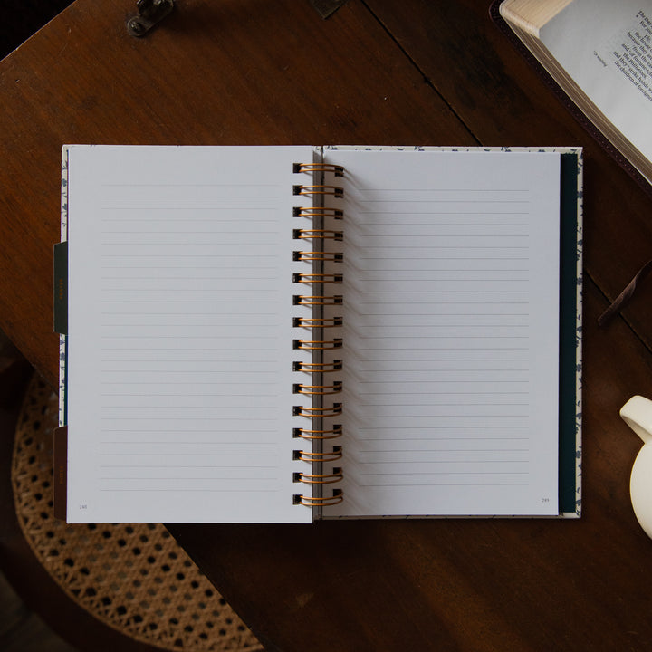 An open spiral-bound lined notebook or planner lies on a dark wooden desk, with part of an open book and a small white object visible around it.