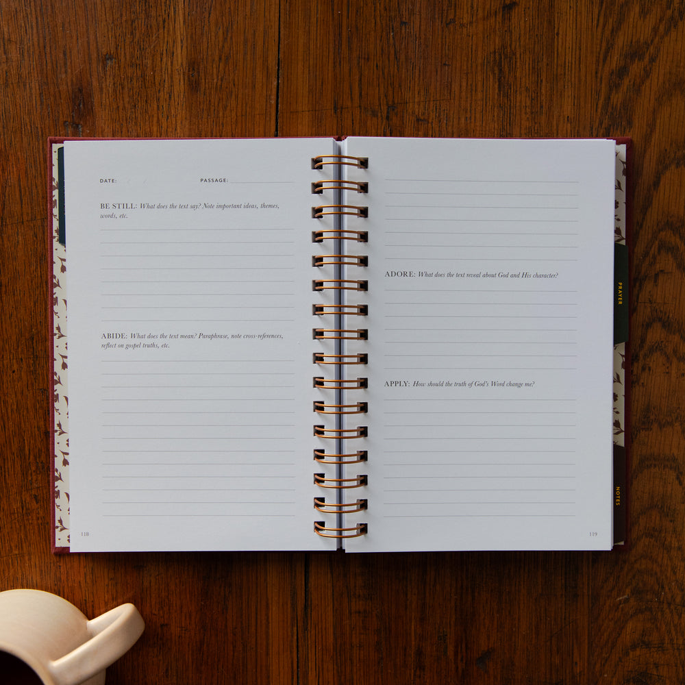 An open spiral-bound journal laid flat on a dark wood tabletop, showing lined pages with printed prompts and a floral-patterned cover edge. A small cream-colored object is partially visible in the lower left corner.