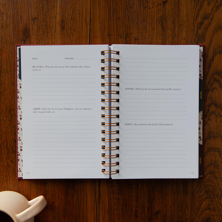 An open spiral-bound journal laid flat on a dark wood tabletop, showing lined pages with printed prompts and a floral-patterned cover edge. A small cream-colored object is partially visible in the lower left corner.