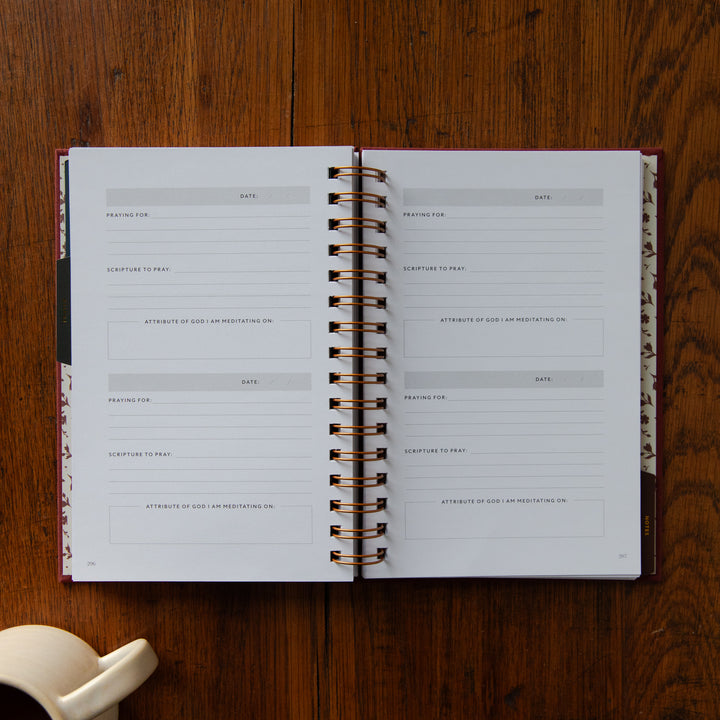 An open spiral-bound planner or journal laid flat on a wooden tabletop, showing blank writing pages with date fields and prayer prompts. A small white ceramic object is partially visible in the lower left corner.