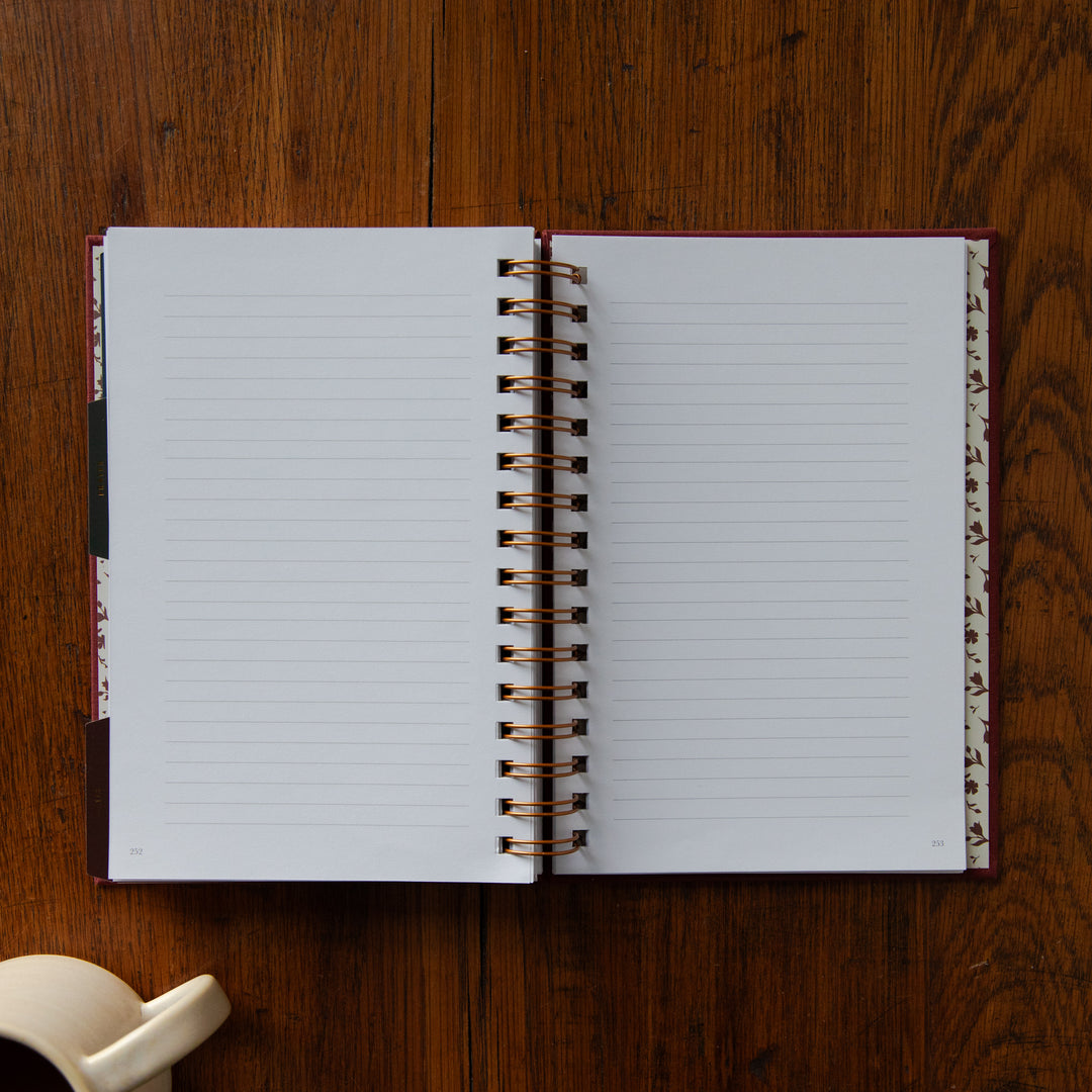An open spiral-bound lined notebook with white pages lies on a dark brown wooden tabletop. A small cream-colored mug is partially visible in the bottom-left corner.