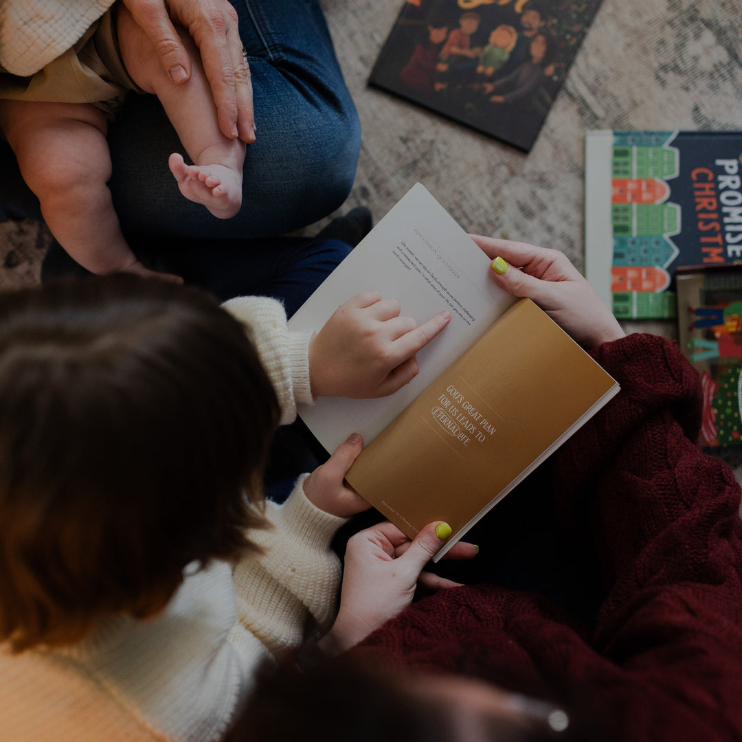 A woman holding open the Advent Adventure devotional while a child points to a sentence in the devotional with various Daily Grace board books laying on the floor
