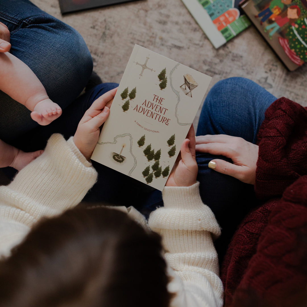A child holding the Advent Adventure Devotional cover next to a woman and a baby