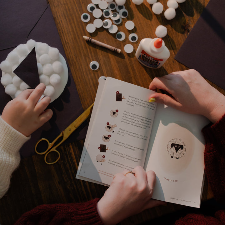 A woman is holding open the Advent Adventure devotional book with a child's hand in a plate of cotton balls
