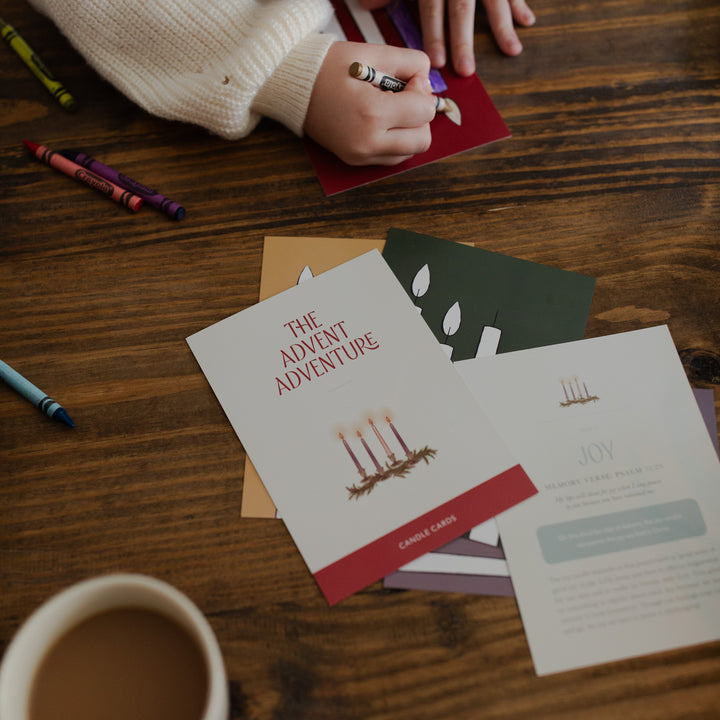 A child's hand coloring a Advent Adventure Card while sitting at a wooden table
