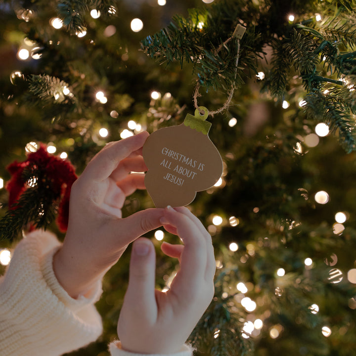 A woman's hand holding an ornament that says Christmas is All About Jesus